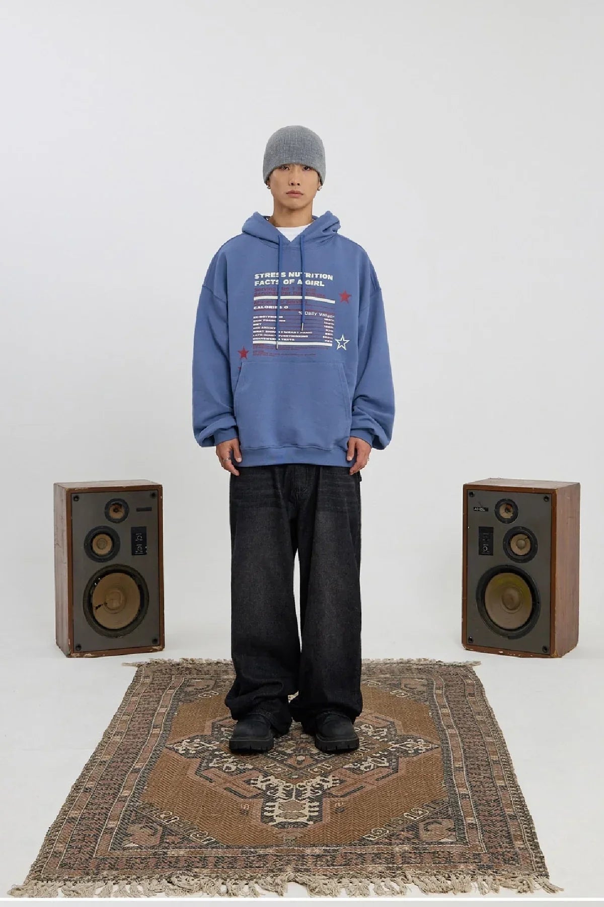 Person wearing dust blue hoodie and gray beanie standing on patterned rug between vintage speakers