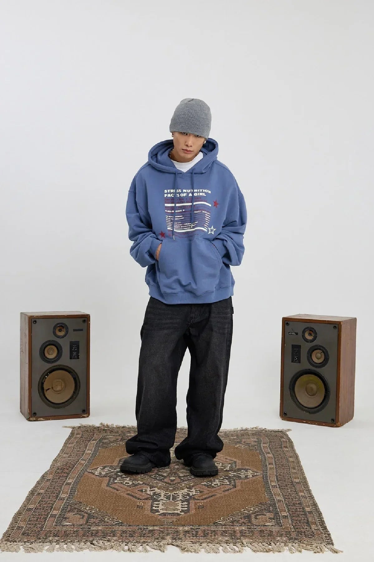 Young man wearing dust blue graphic hoodie, gray beanie, and black pants standing on a patterned rug between vintage speakers