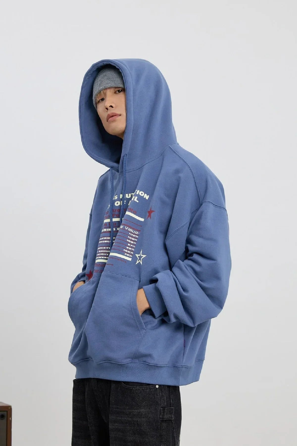 Man wearing dust blue graphic hoodie and gray beanie with hands in pockets against white background