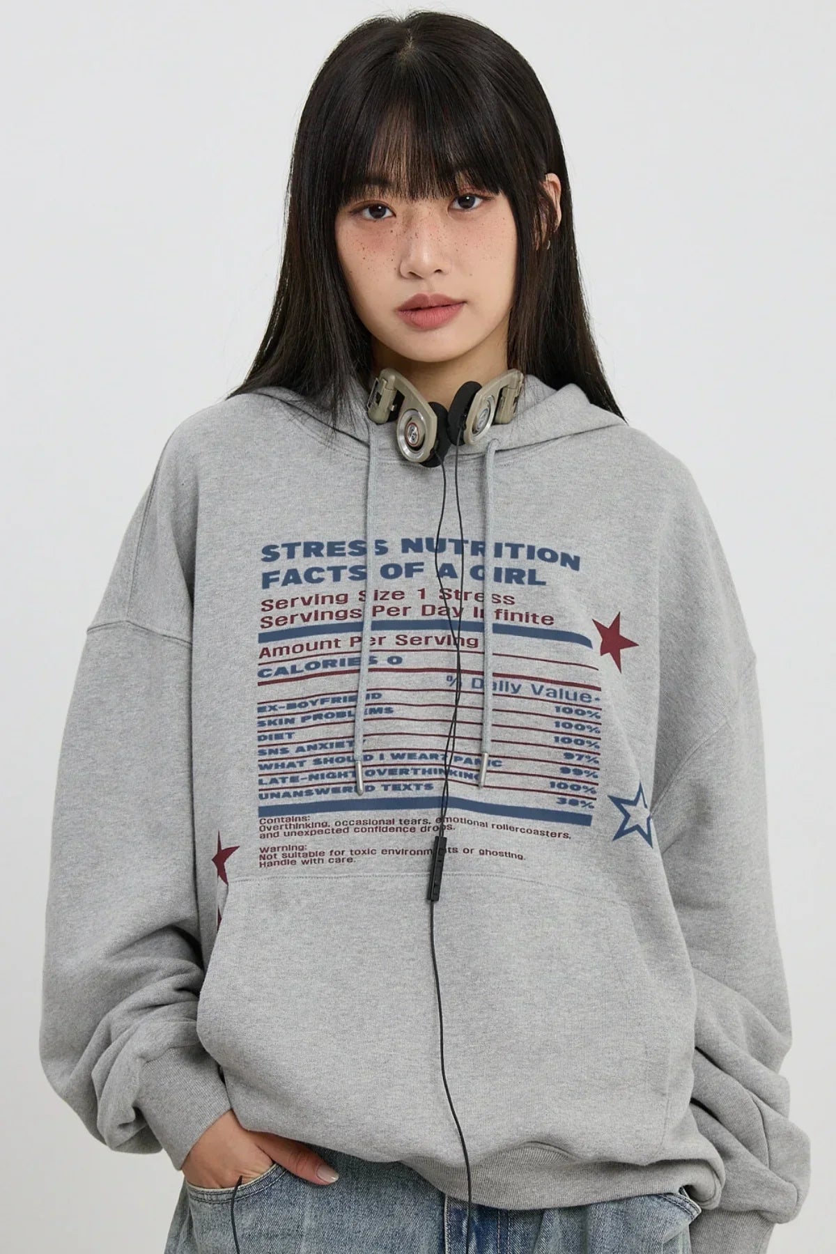 Young woman wearing a grey hoodie with stress nutrition facts print and headphones around neck