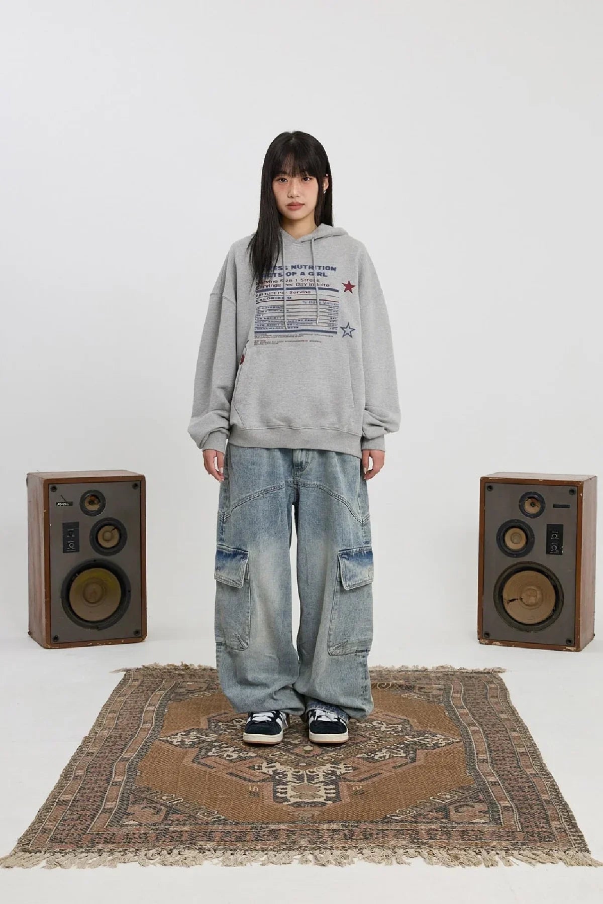 Young woman wearing grey nutritional info hoodie and baggy jeans standing on patterned rug with speakers