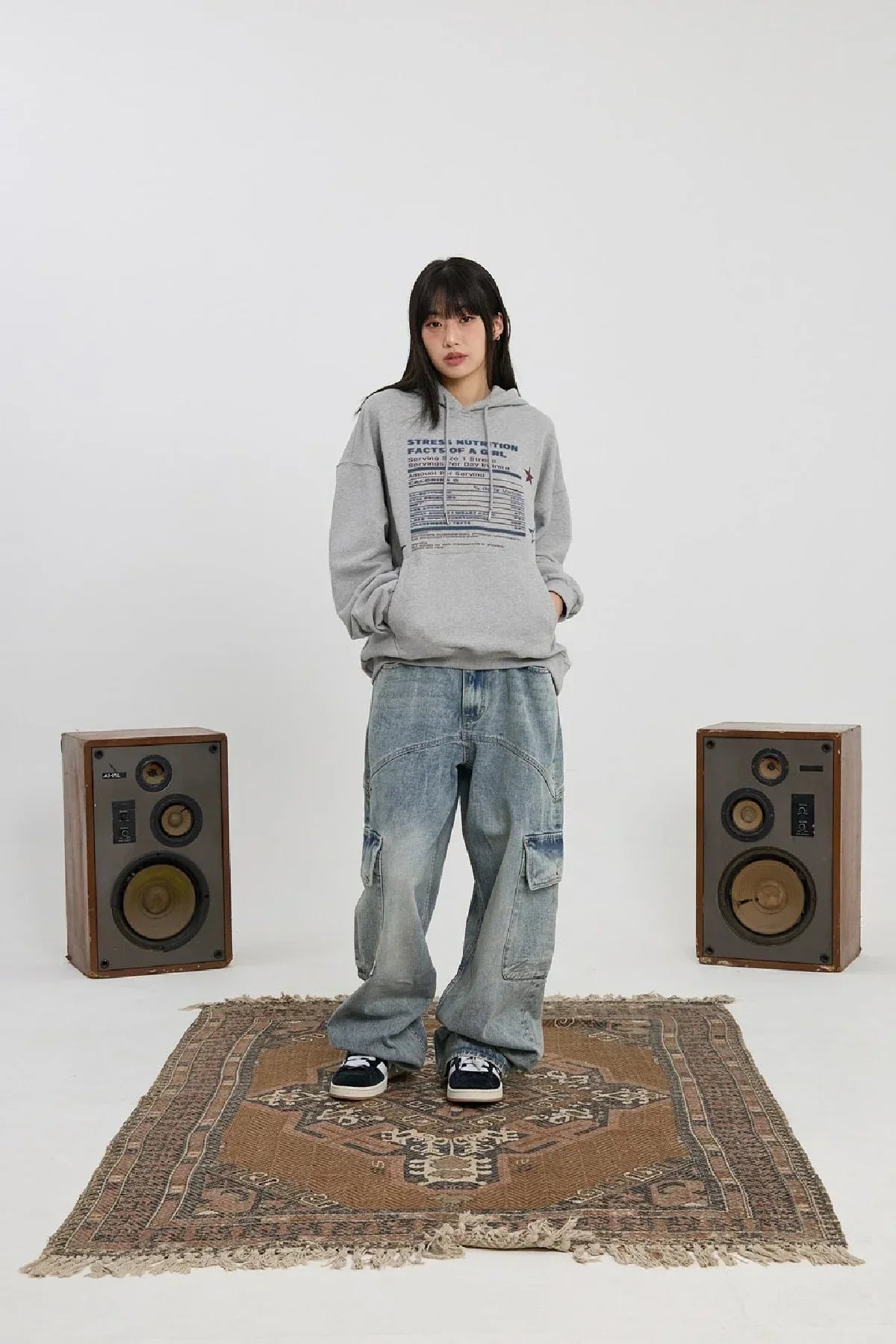 Young woman wearing grey hoodie and baggy jeans standing on patterned rug between vintage speakers