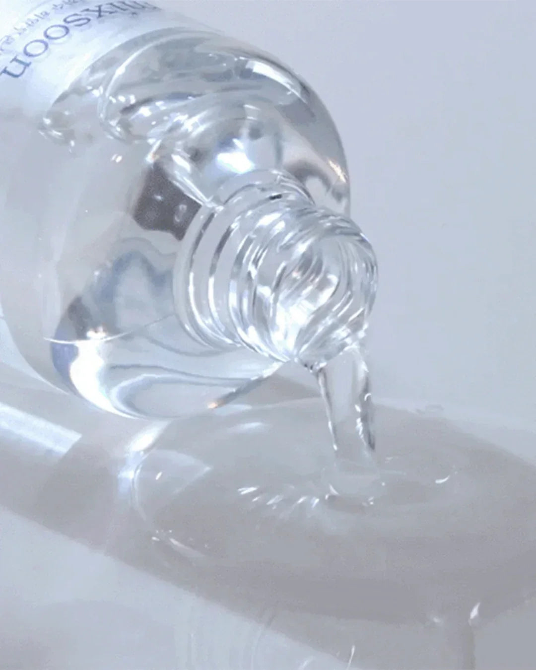 Close-up of clear hyaluronic acid serum pouring from a clear bottle on white surface