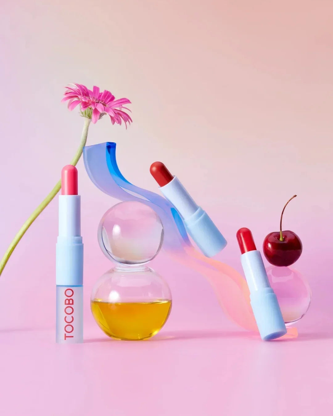 Three tinted lip balms in white tubes with pink and red shades, accompanied by a pink flower, cherry, and glass spheres on pink background