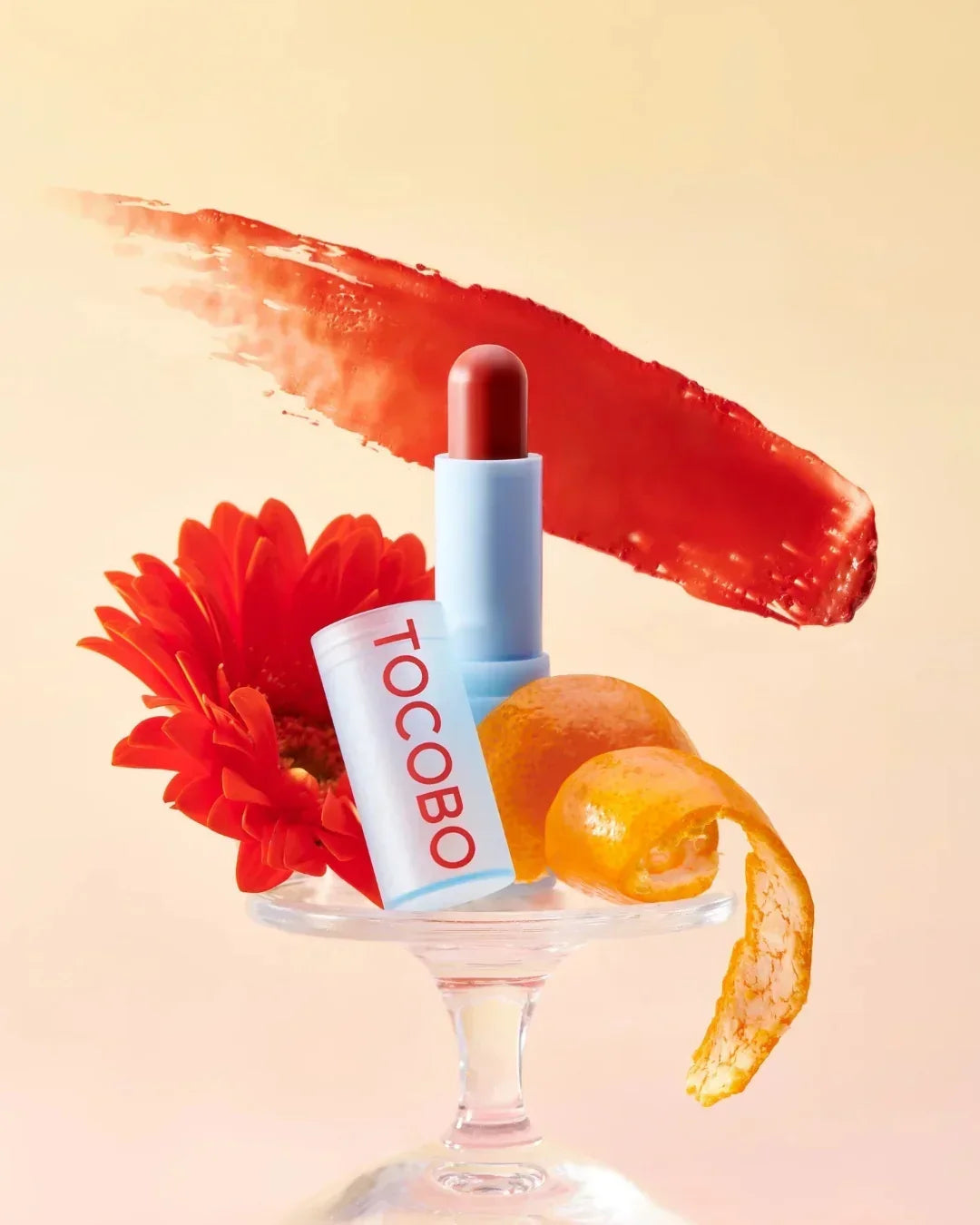 TOCOBO tinted lip balm on glass stand with red flower, orange peel, and red color smear background