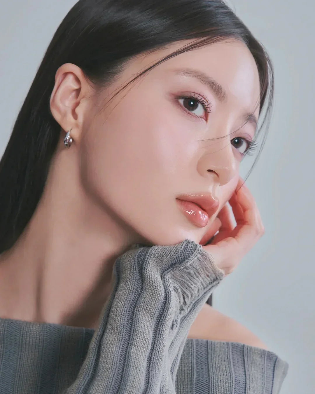Close-up of woman with glossy lips, natural makeup, sleek black hair, and gray knit sweater against gray background
