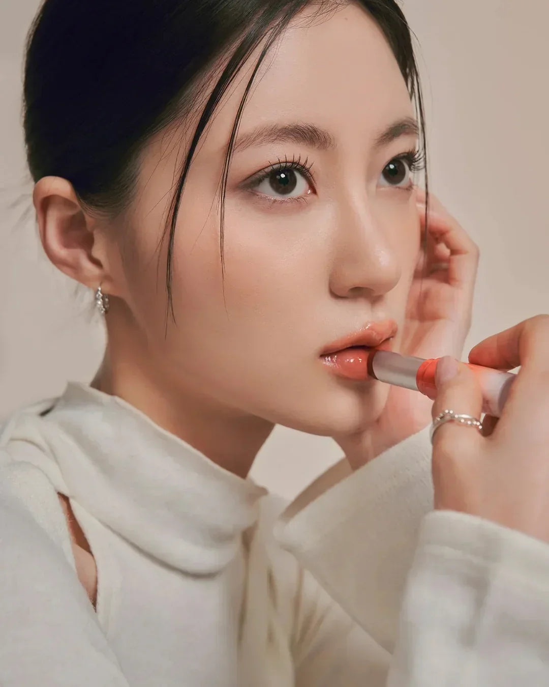 Close-up of a woman applying glossy peach lip balm with a silver tube against a neutral background
