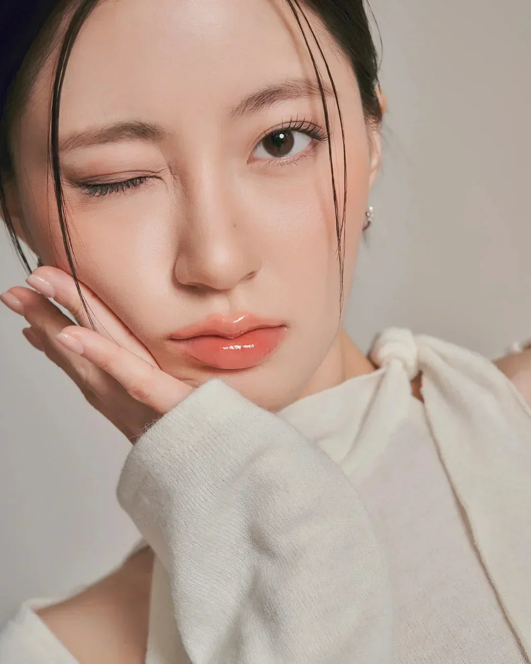 Close-up of woman with glossy peach lips, natural makeup, hand on face, and cream knit top