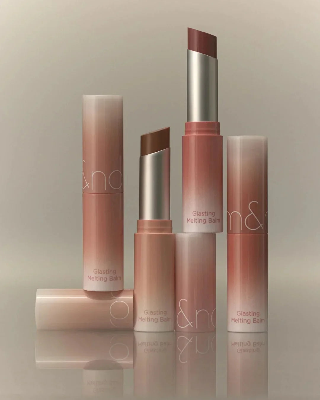 Three Glasting Melting Balm lipsticks in pink and brown shades with gradient packaging on reflective surface