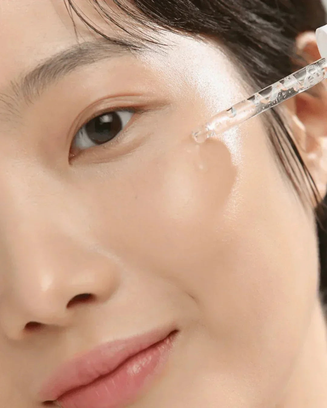 Close-up of woman applying clear serum to glowing, smooth cheek with dropper