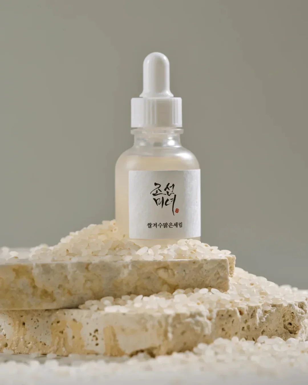 Glass dropper bottle serum on stone blocks surrounded by white rice grains, natural skincare essence