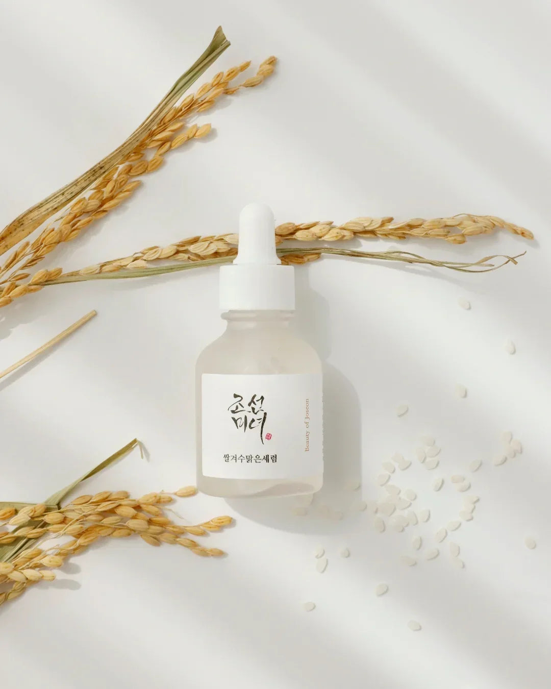 Frosted glass serum bottle with white dropper on white surface surrounded by dried rice stalks and scattered rice grains