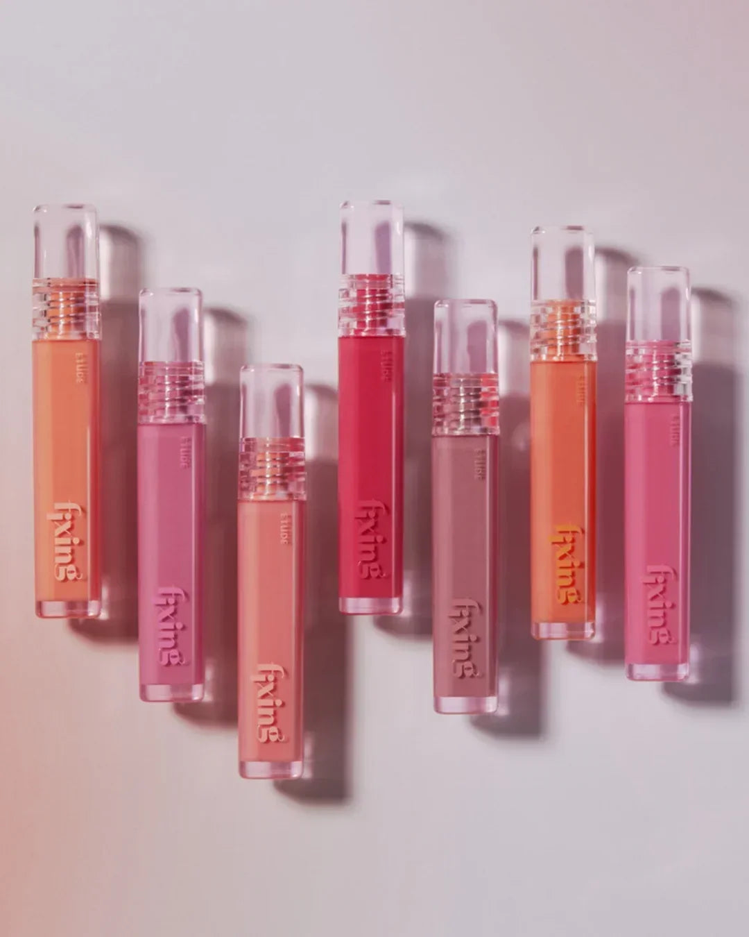 Seven colorful Fixing tint lipsticks arranged on a light pink background for makeup and beauty