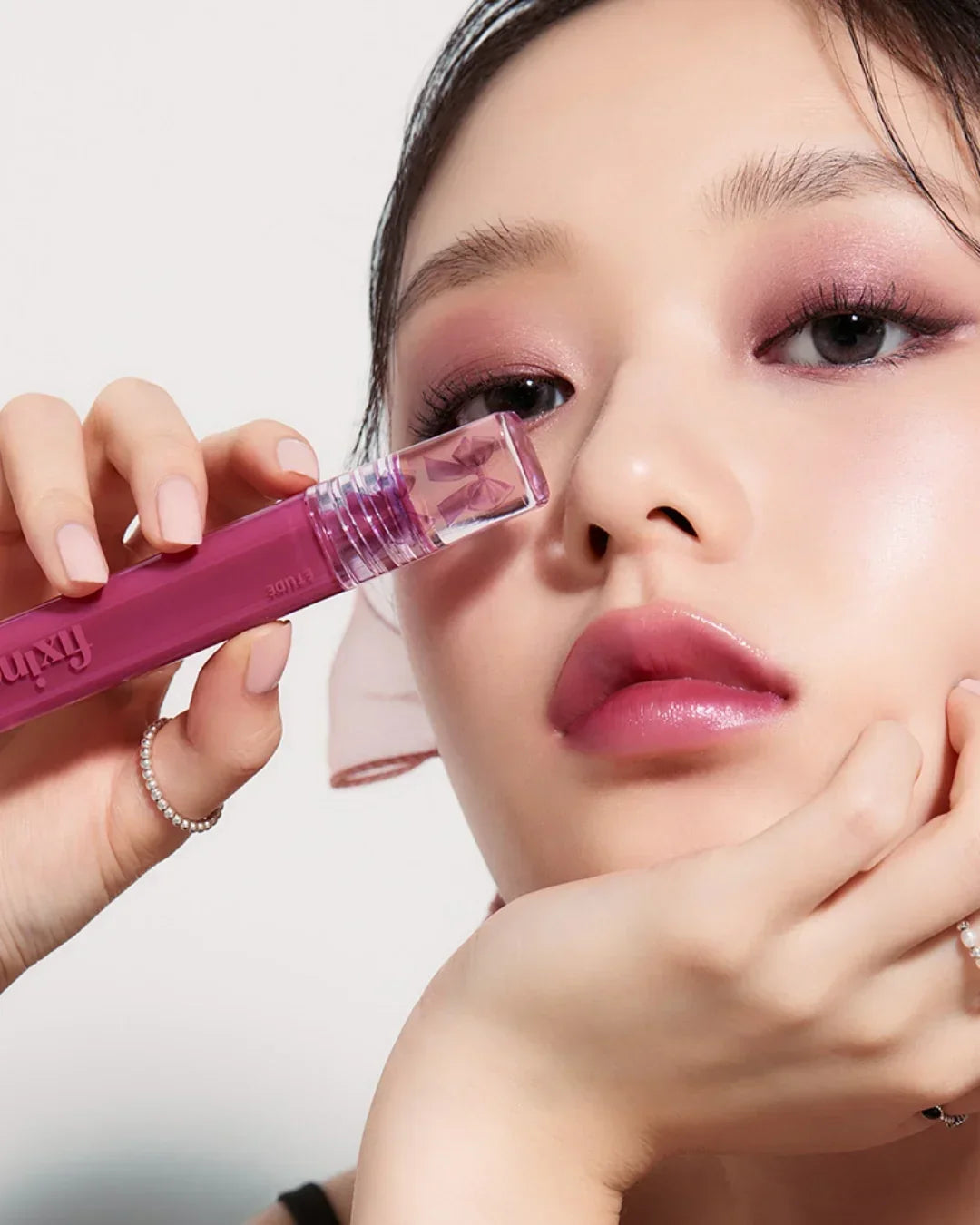 Close-up of woman with pink eye shadow and glossy lips holding a purple Fixing Tint makeup product