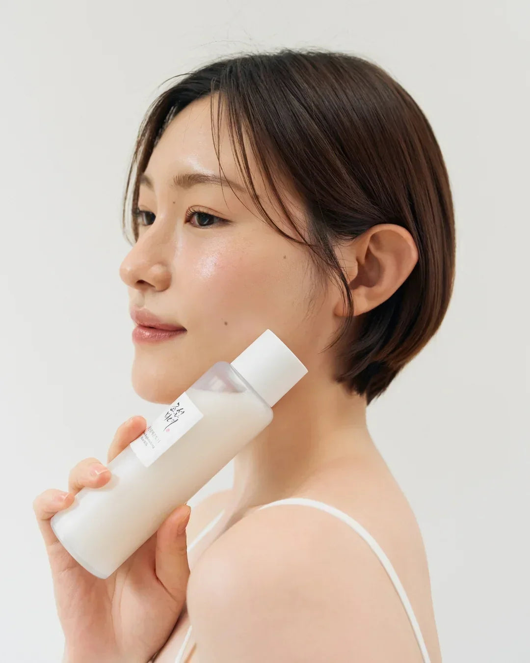 Close-up of woman with glowing skin holding a white skincare bottle near her face, minimal background