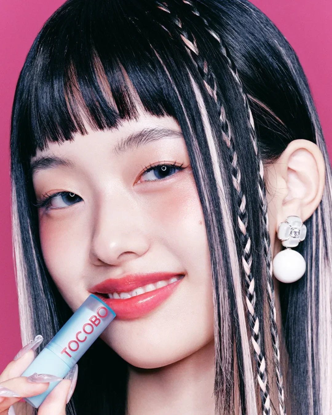Young woman with braided black and blonde hair applying Tocobo lip balm against pink background