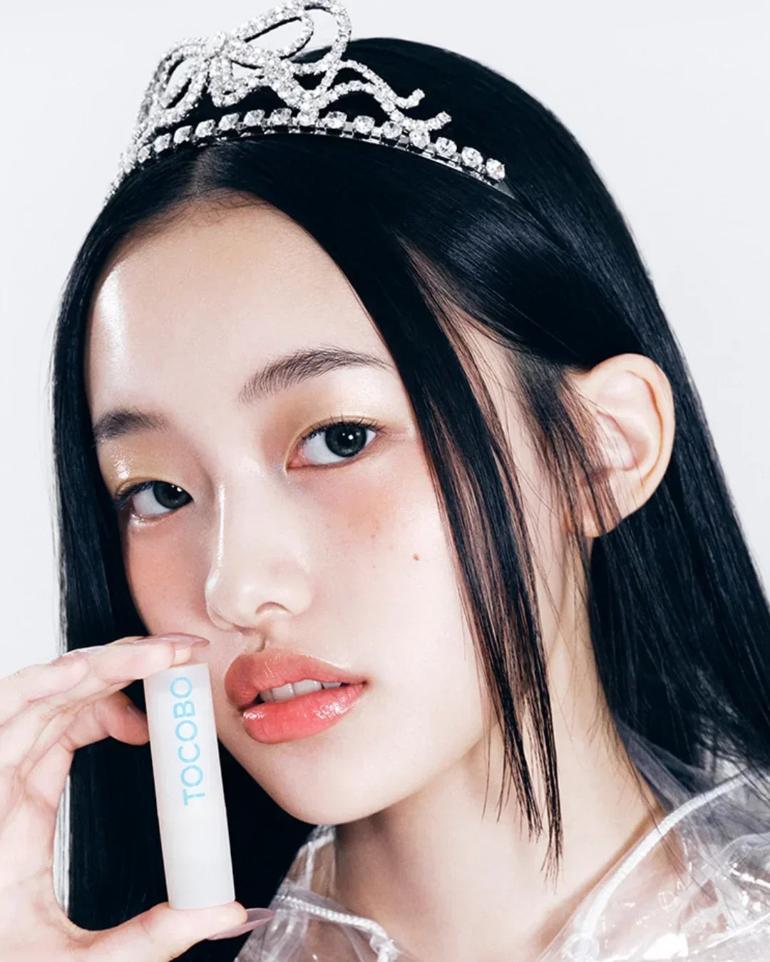 Young woman wearing a rhinestone tiara holding Tocobo lip balm close to her face with glossy lips