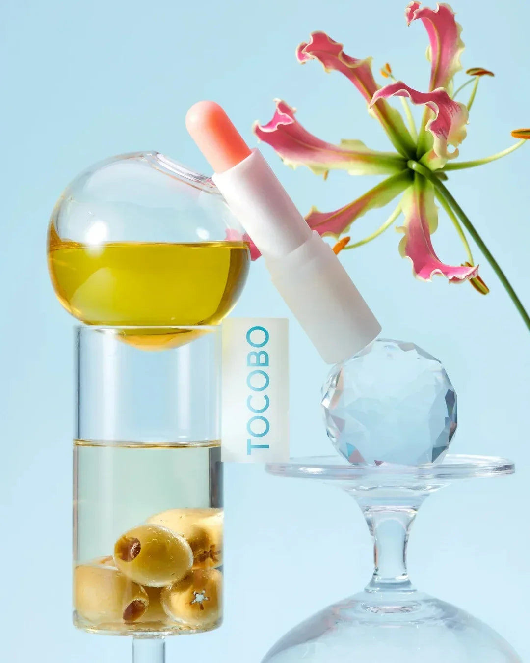 TOCOBO lip balm with peach color, flower, olives in glass containers, and crystal ball on light blue background