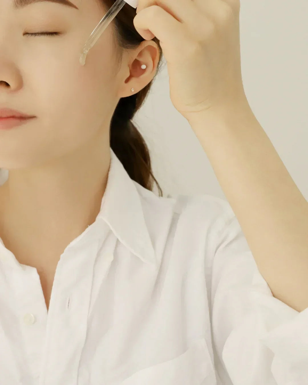 Woman wearing white shirt applying clear facial serum with dropper on cheek skin care