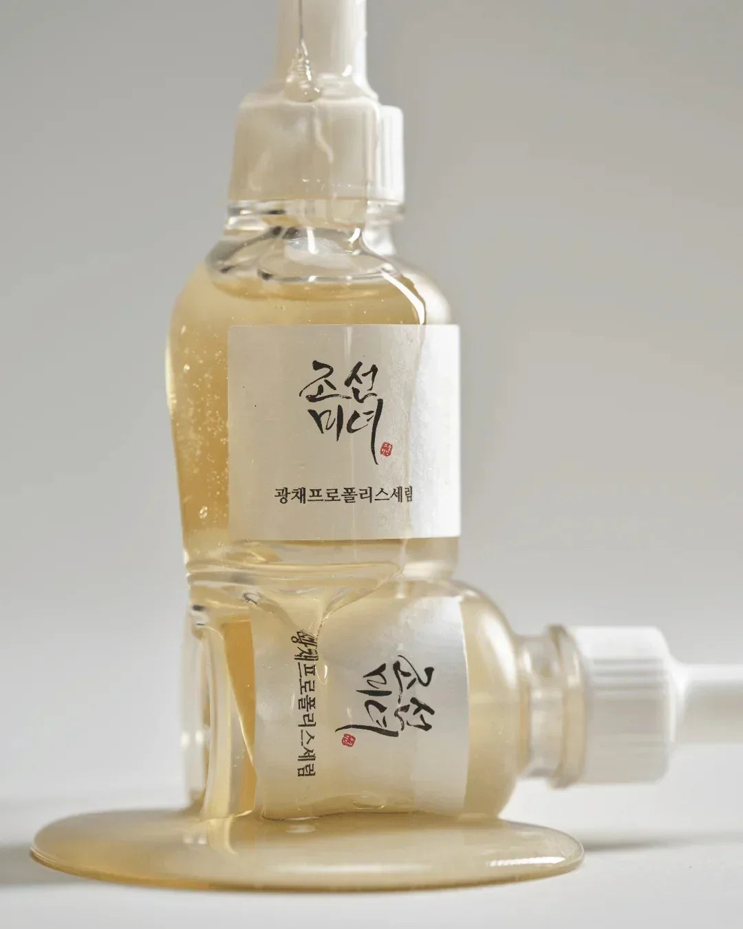 Two dropper bottles of Korean propolis serum with golden liquid spilling on white surface