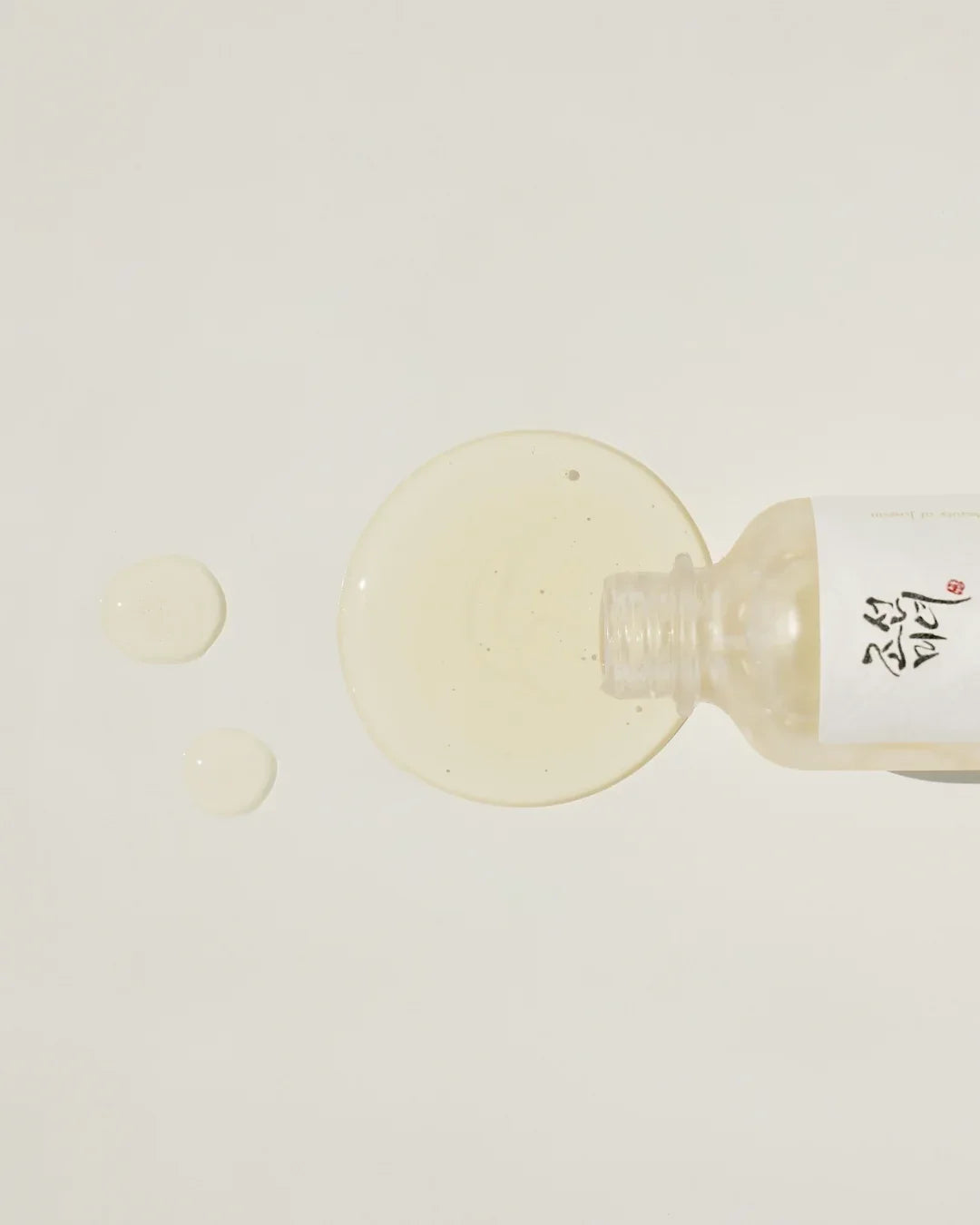 Open bottle spilling clear light serum drops on white surface, skincare product