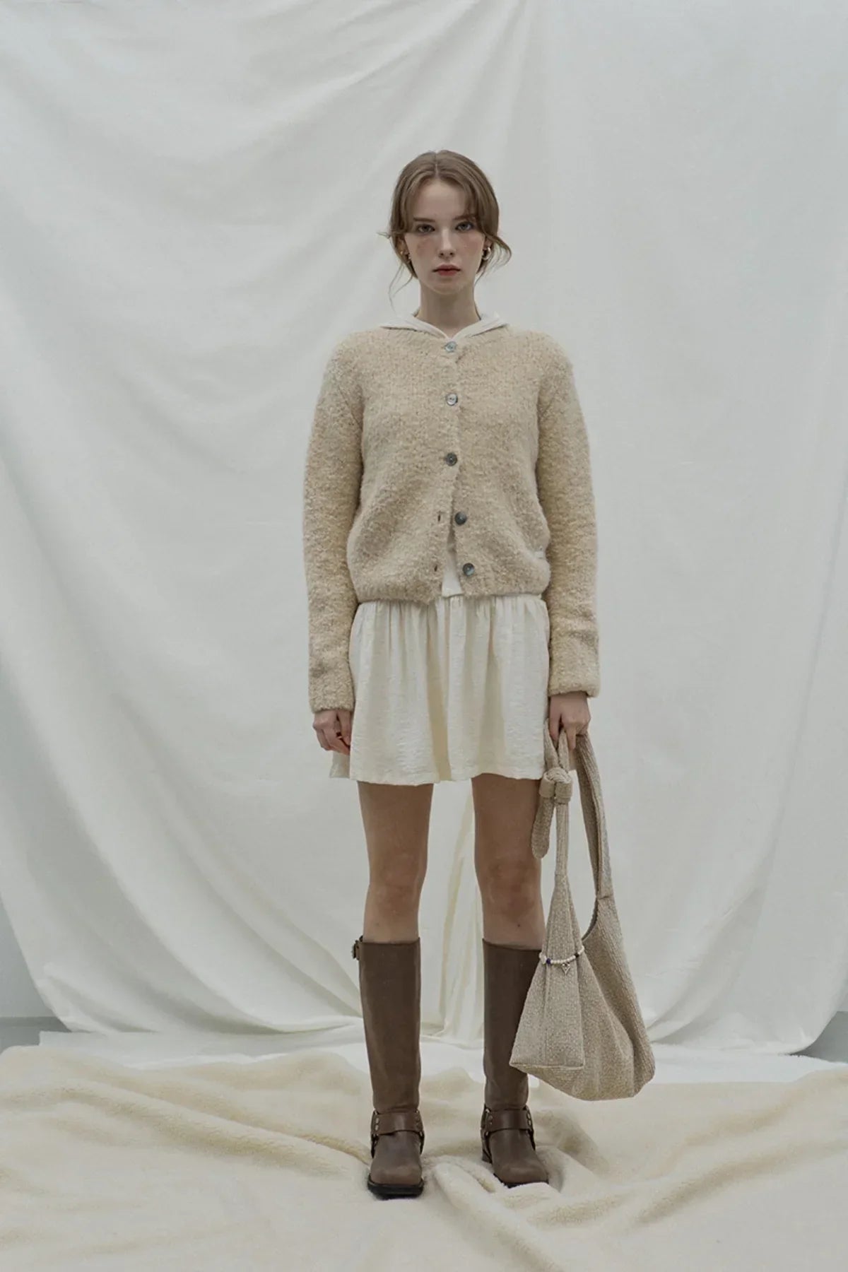 Young woman wearing beige grandma cardigan, white dress, brown boots, holding matching beige bag