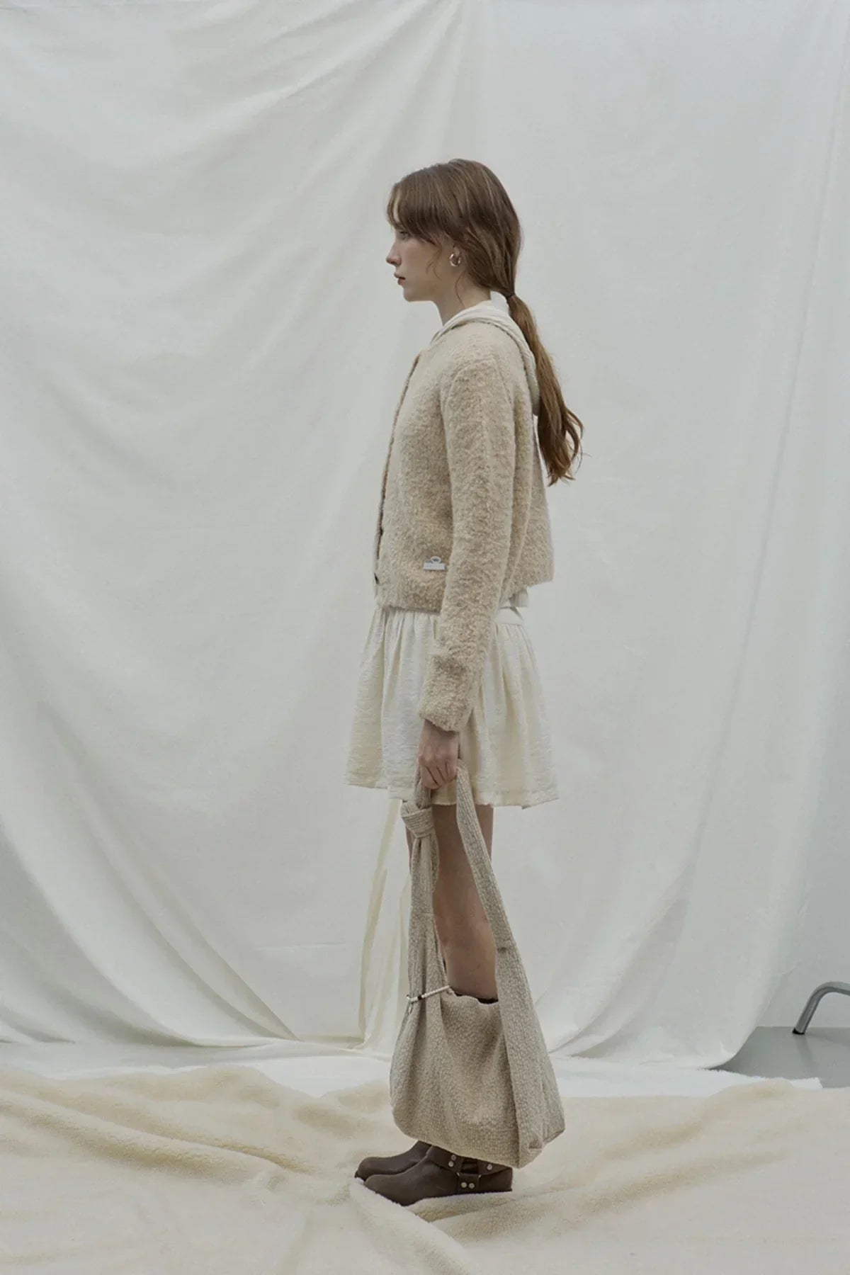 Side view of woman wearing beige grandma cardigan and white dress, holding matching knit bag in minimalist studio