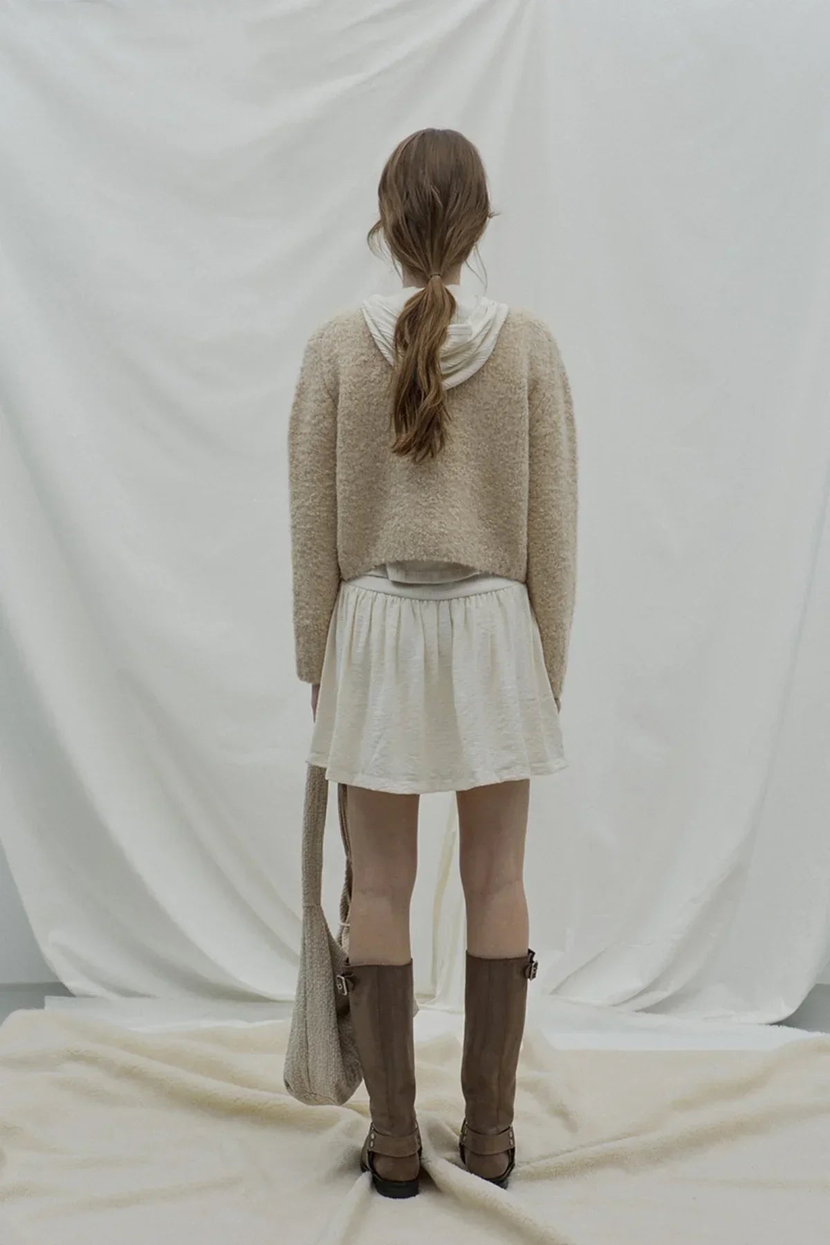 Woman wearing beige fuzzy cardigan with white skirt and brown boots, standing on beige fabric backdrop