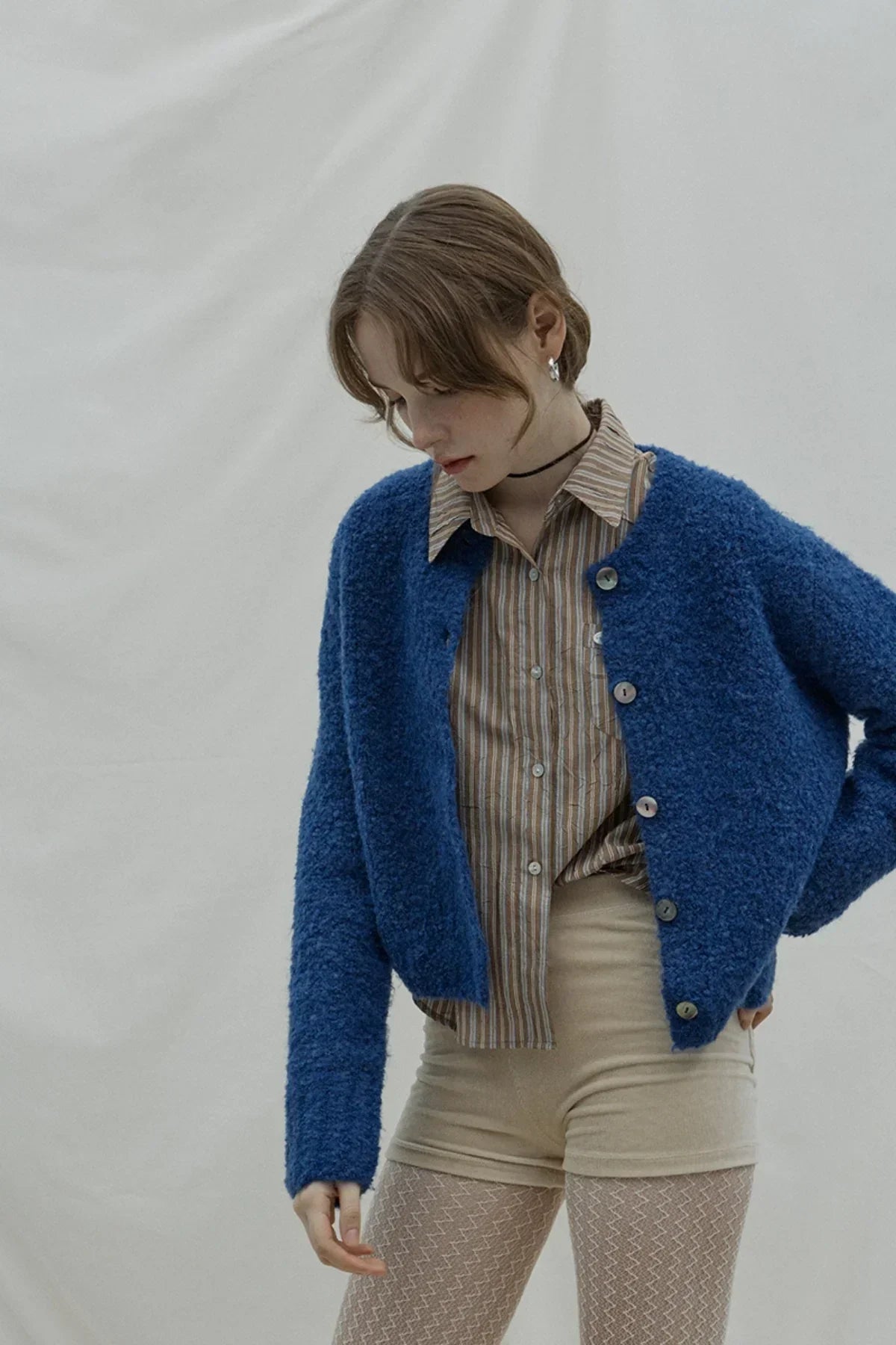 Young woman wearing a dark blue buttoned cardigan, striped shirt, and beige shorts against a neutral background