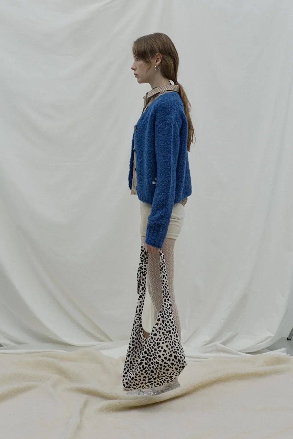 Side view of woman wearing dark blue grandma cardigan, striped shirt, white shorts and holding spotted tote bag