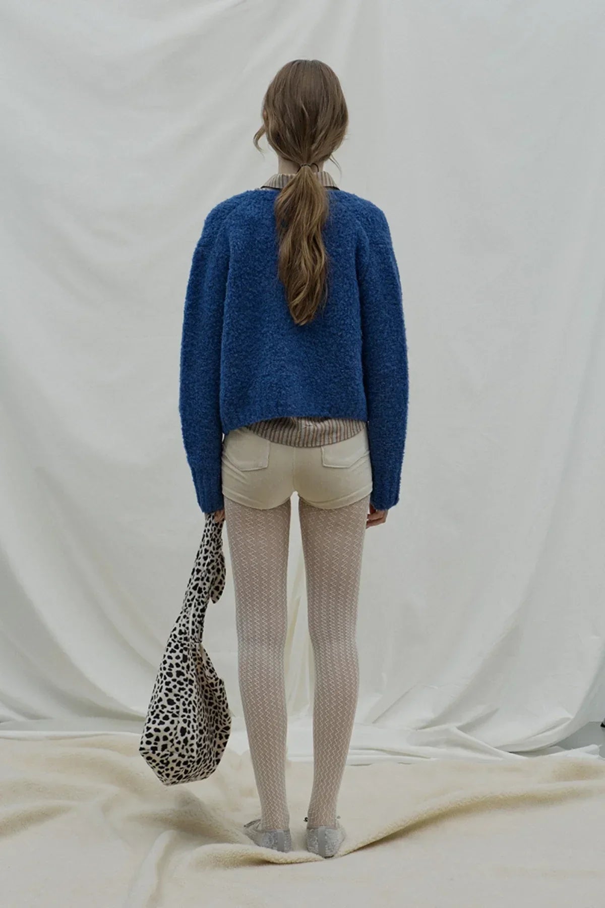 Back view of woman wearing dark blue fuzzy cardigan, beige shorts, patterned tights, holding leopard print bag