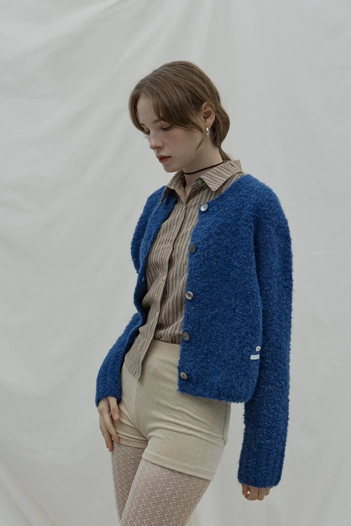 Woman wearing a dark blue textured cardigan over beige shorts and patterned tights on plain background