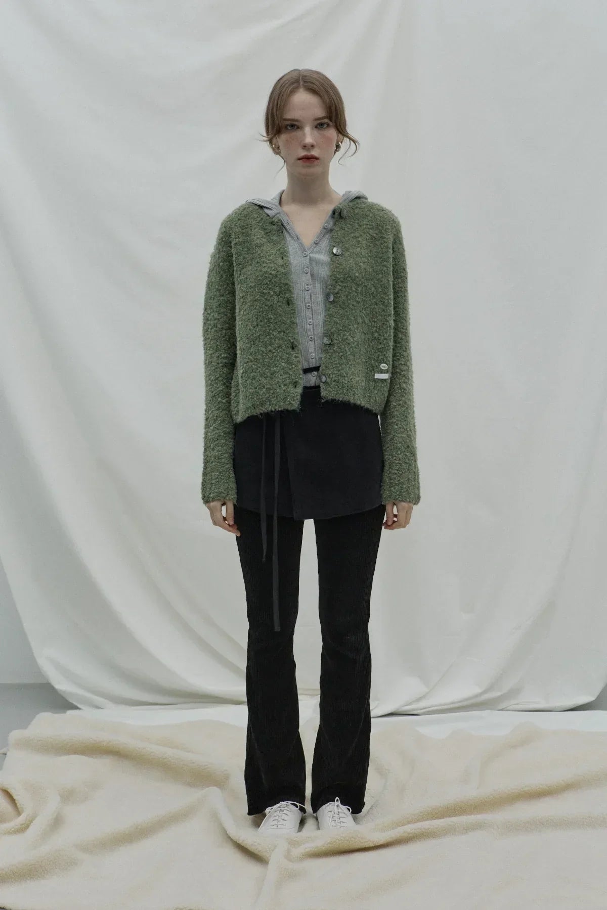 Woman wearing green fluffy cardigan, grey hoodie, black pants, and white sneakers in studio setting