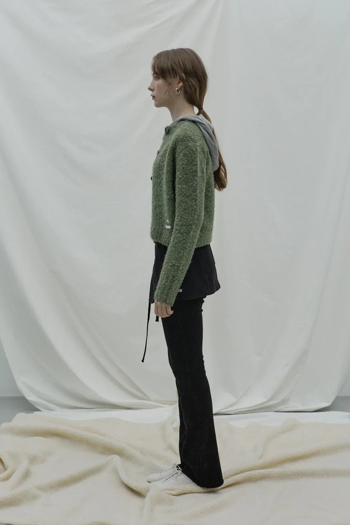 Side profile of woman wearing green fuzzy grandma cardigan over gray hoodie with black pants and white shoes
