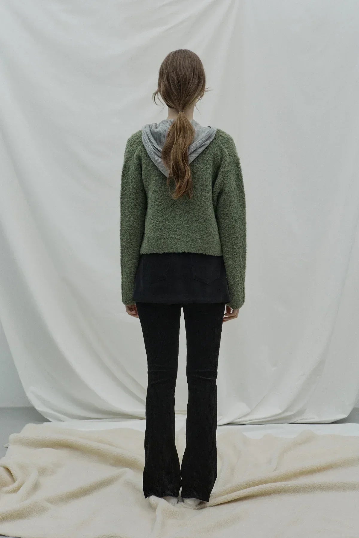 Woman wearing green textured cardigan with gray hood and black pants standing against white backdrop