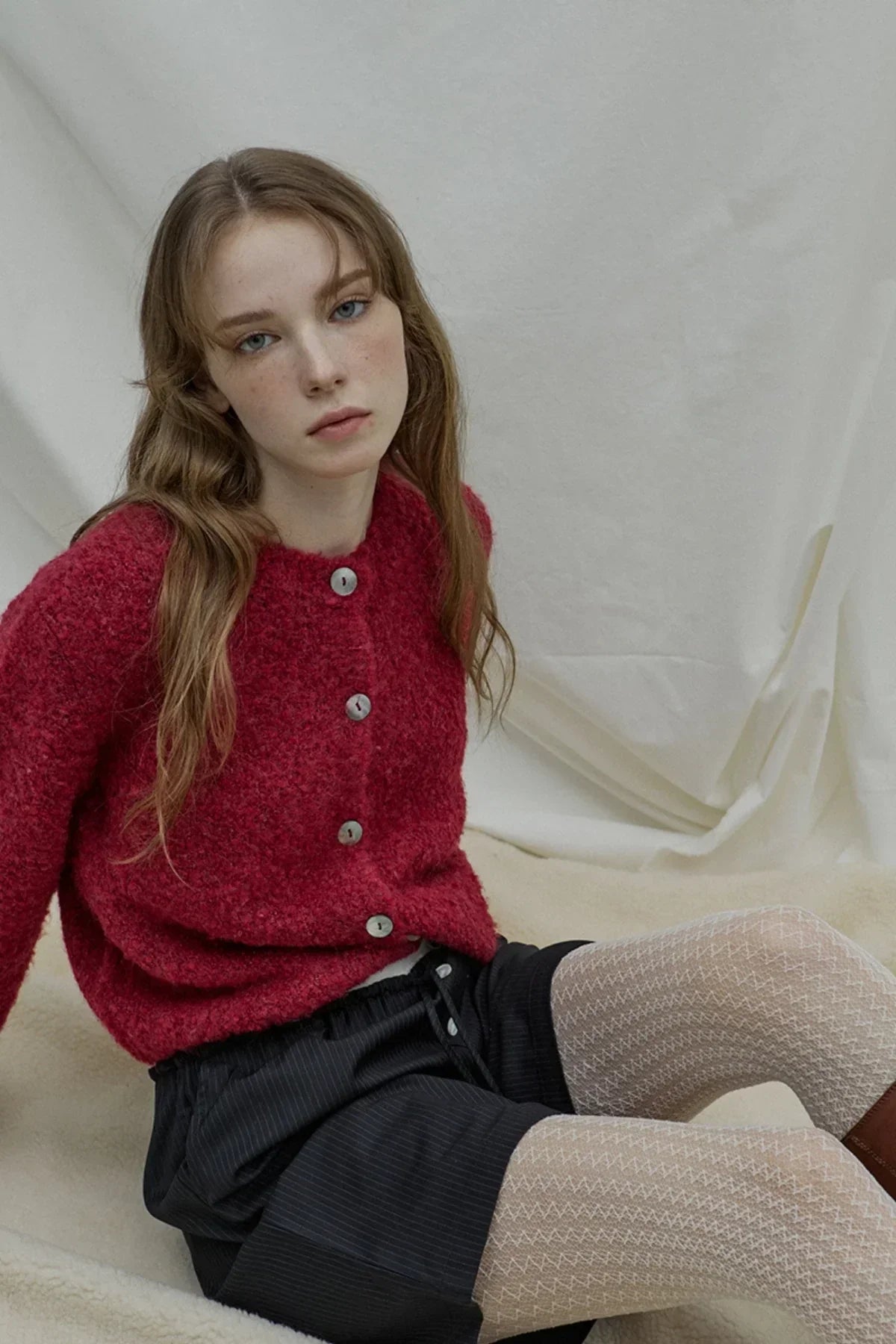 Young woman wearing a textured red cardigan with buttons, black shorts, and patterned tights sitting against a neutral backdrop
