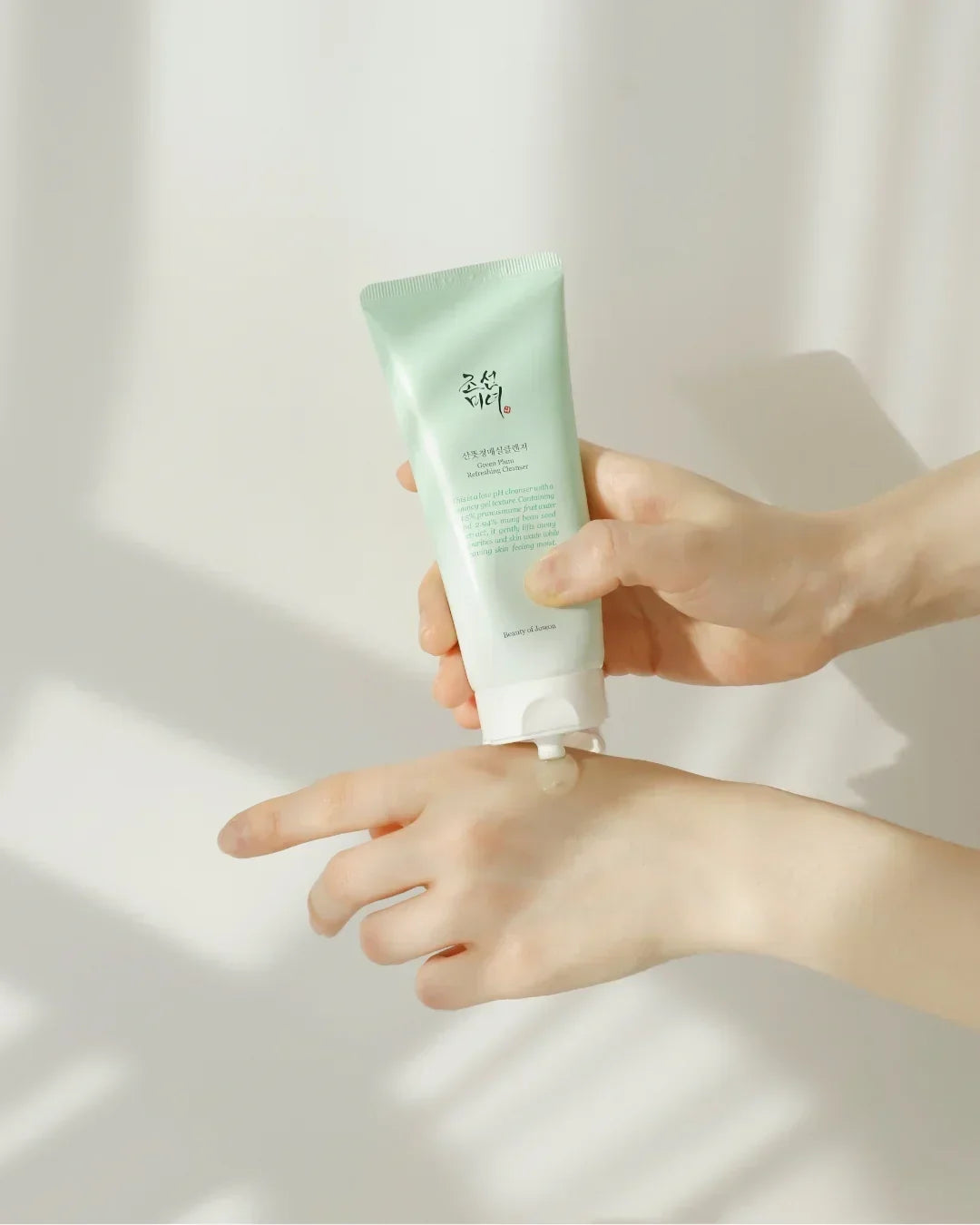 Hand squeezing green plum refreshing cleanser gel from pastel green tube onto wrist in soft natural light