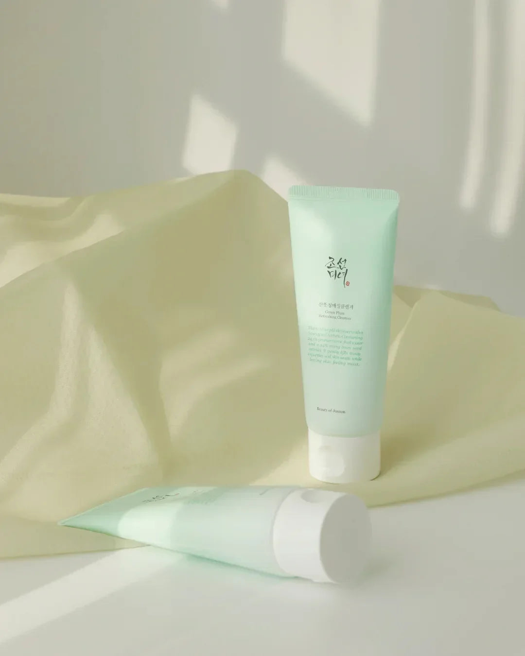 Two pastel green tubes of Green Plum Refreshing Cleanser on soft beige fabric under natural light