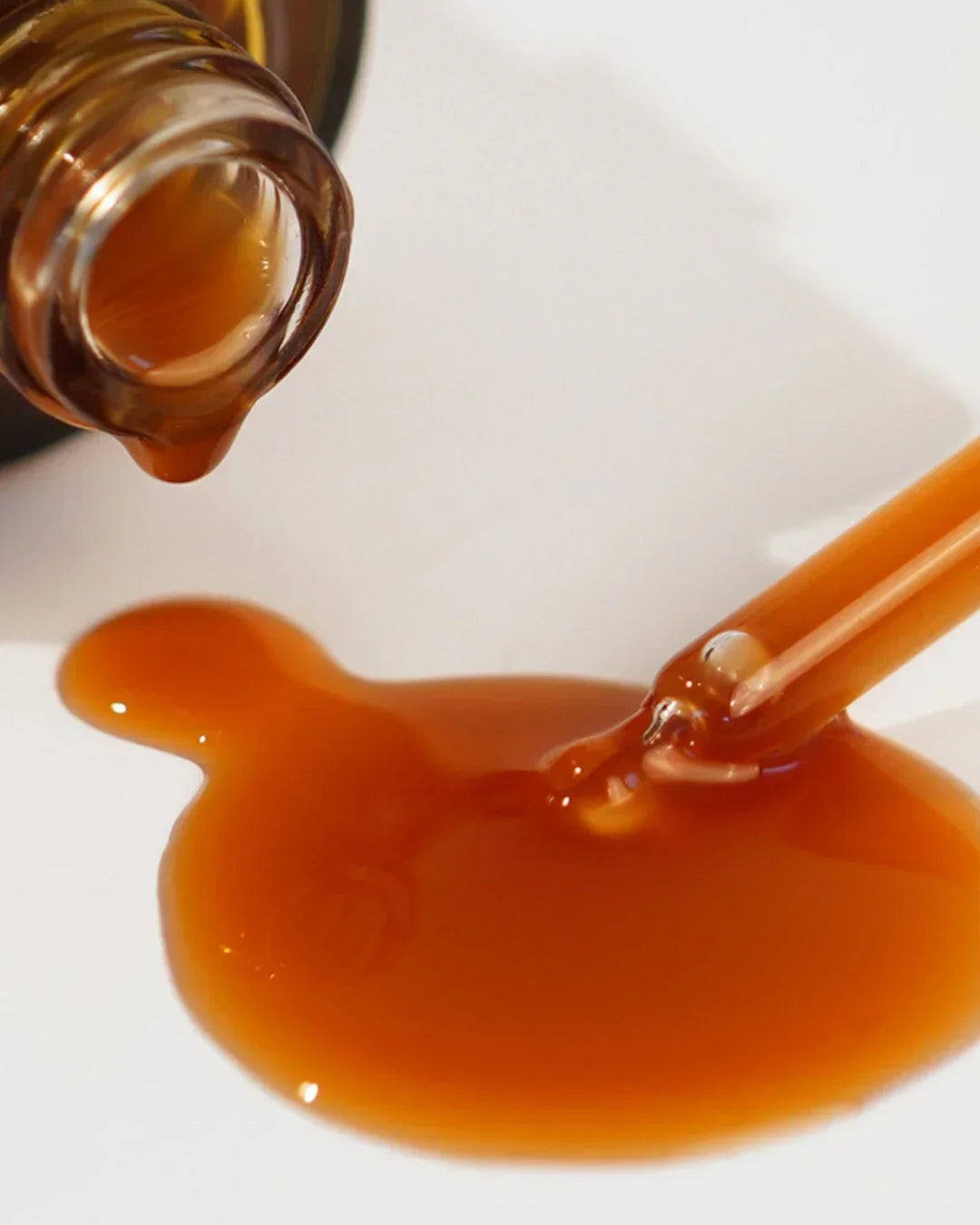 Close-up of amber serum dripping from bottle and dropper on white surface