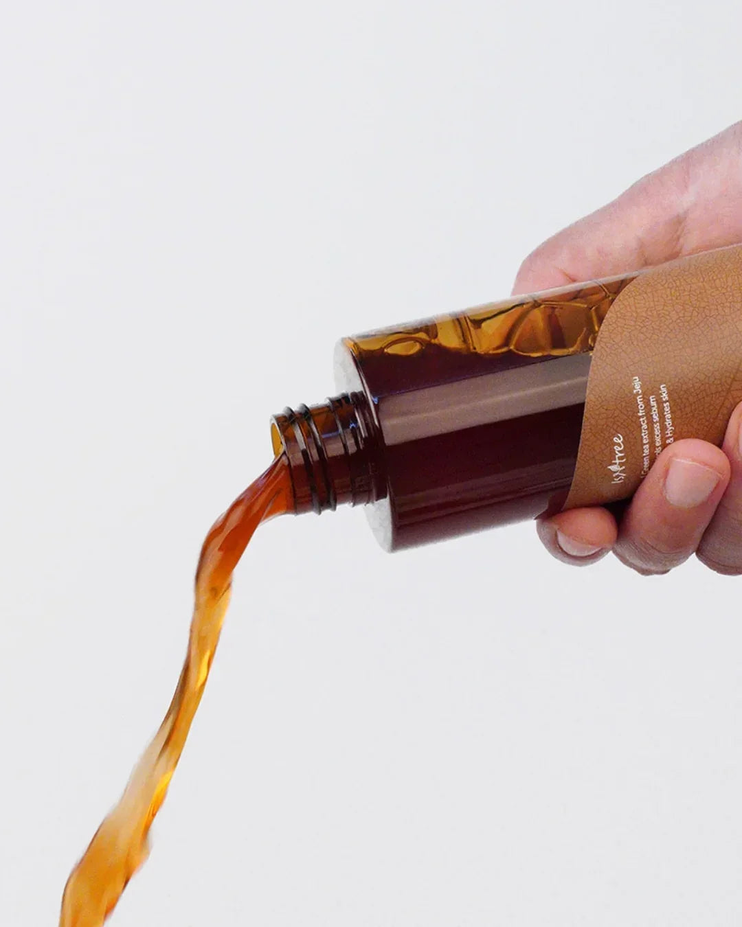 Hand pouring amber liquid from a brown glass skincare bottle with brown label on white background