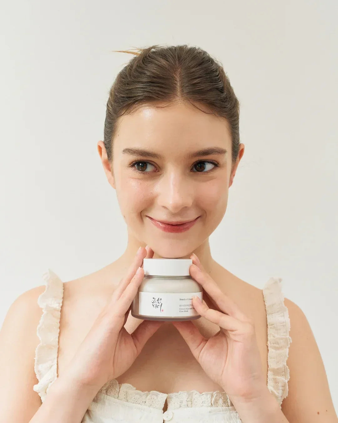 Smiling young woman in white dress holding jar of Ground Rice and Honey Glow Mask skincare