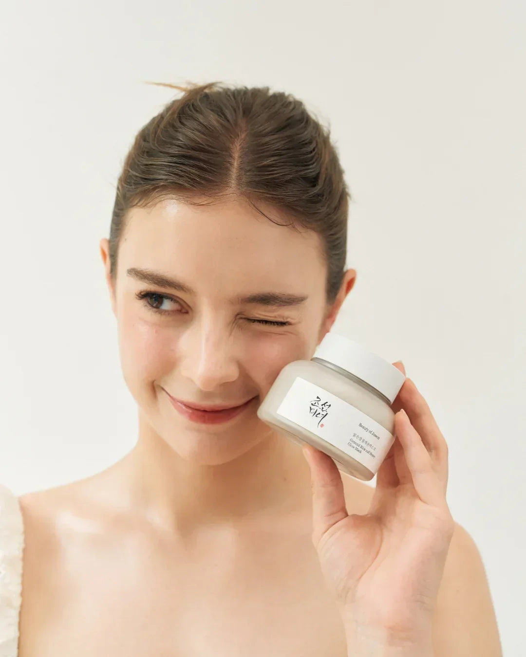 Smiling woman with clear skin holding Ground Rice and Honey Glow Mask jar near face