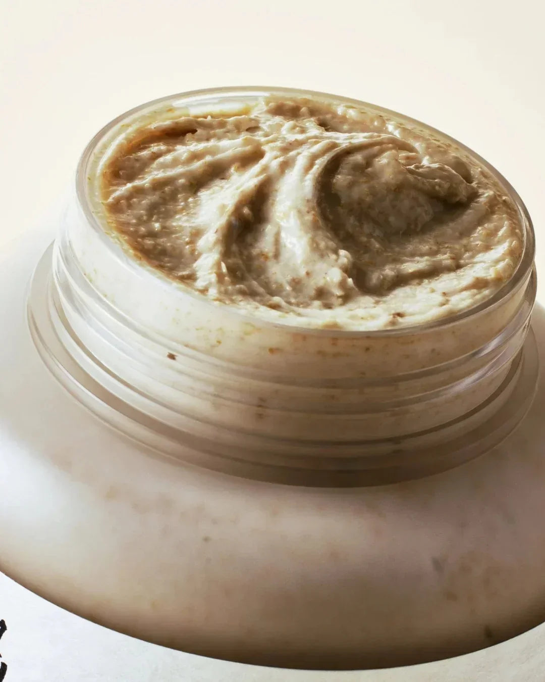 Close-up of a creamy, grainy textured rice and honey glow mask in a clear jar on a light background