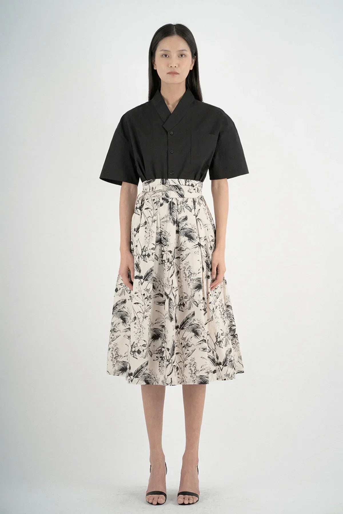Woman wearing a black short-sleeved shirt paired with a high-waisted beige floral midi skirt and black heels on white background