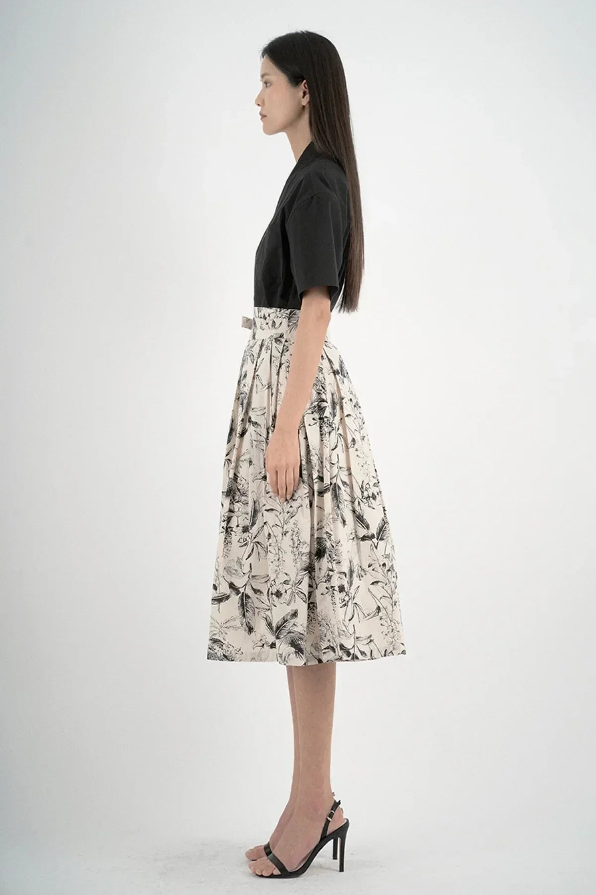 Profile of woman in black short-sleeve shirt, floral midi skirt, and black heeled sandals on white background