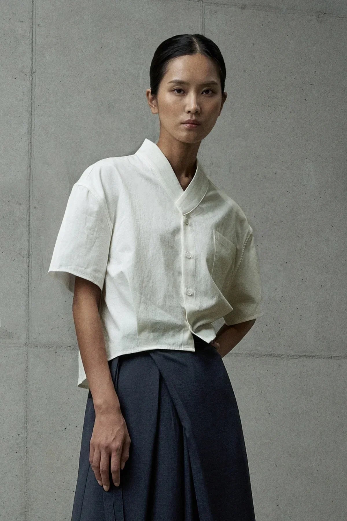 Model wearing ivory short-sleeved button-up shirt with mandarin collar and dark pleated skirt
