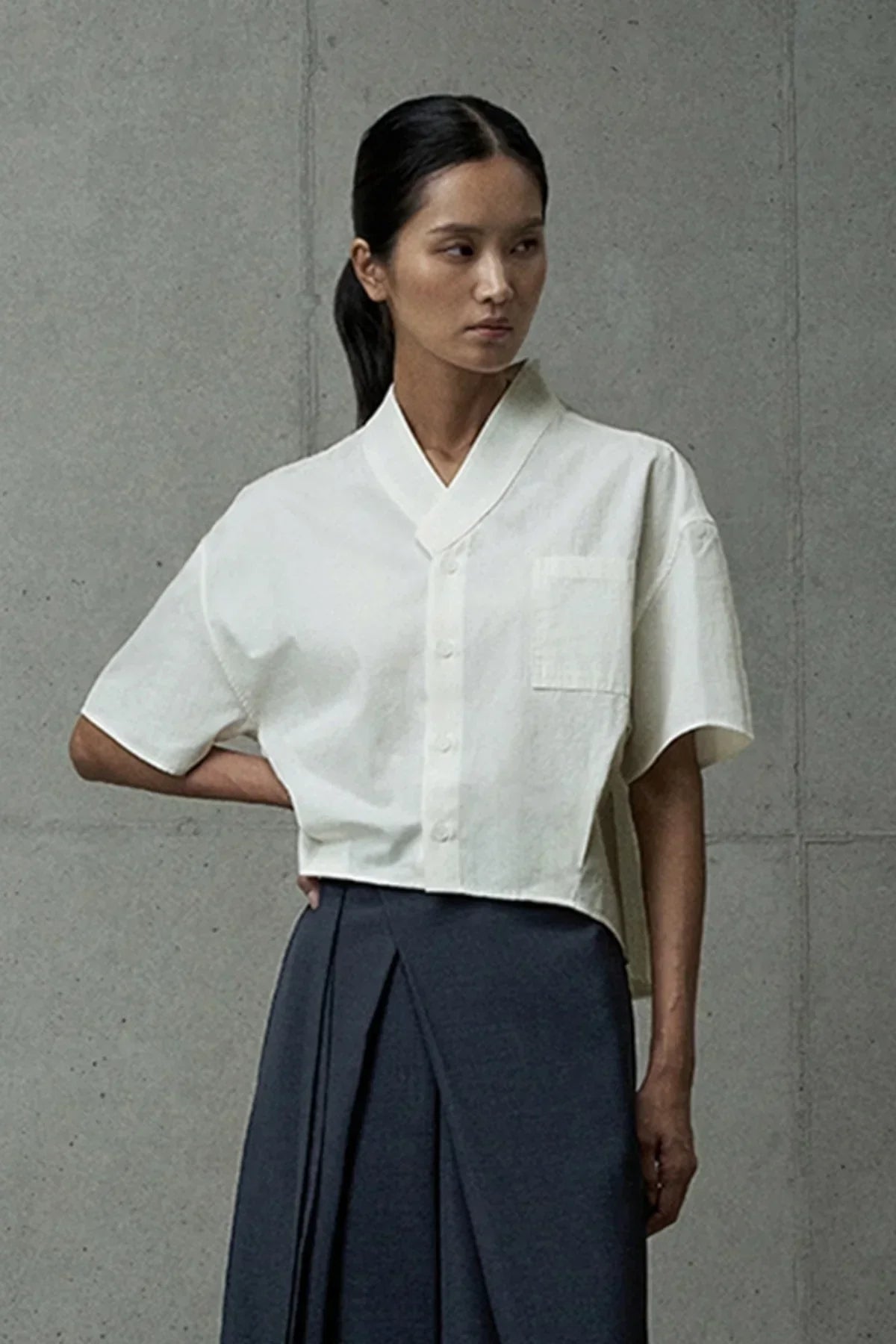 Woman wearing an ivory short-sleeved button-up shirt with a chest pocket and dark pleated skirt
