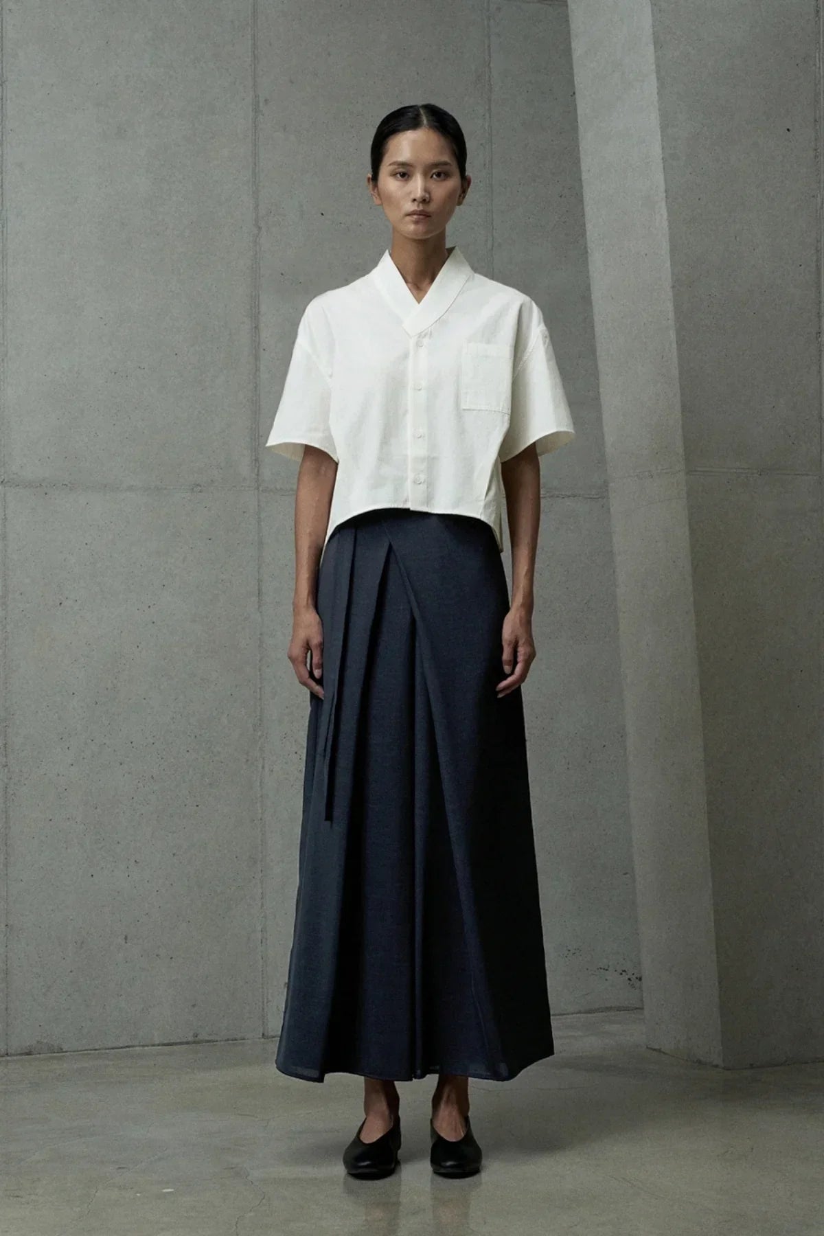 Woman wearing a white short-sleeved cropped shirt and a high-waisted dark wrap skirt in minimalist concrete interior