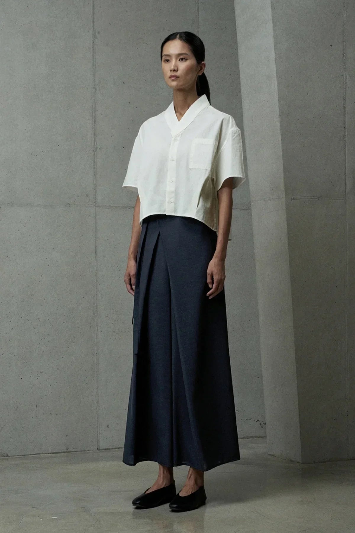 Woman in ivory short-sleeved button-up shirt and dark wide-leg pants standing against concrete wall