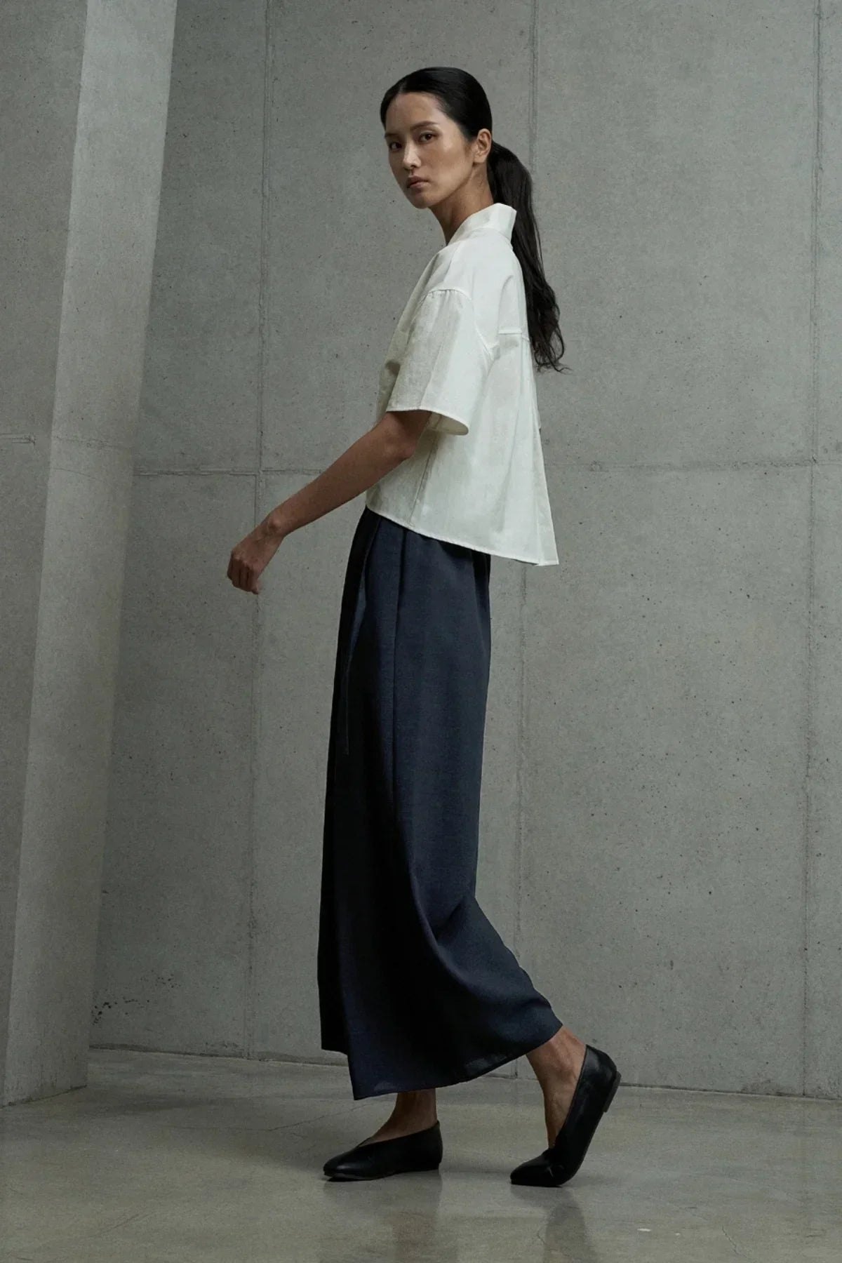 Woman modeling ivory short-sleeved shirt with black wide-leg pants and flats against concrete wall